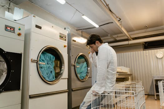 An adult man sits patiently in a laundry facility next to industrial washers.